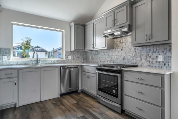 Picture of interior "Ideal Home" kitchen with grey cabinets and stainless steel appliances, with large window above the sink. 