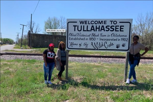 Charity Marcus, Cymone Davis and Vanessa Morrison next to sign that says "Welcome to Tullahassee, Oldest Historical Black Town in Oklahoma, Home of the Wildcats".