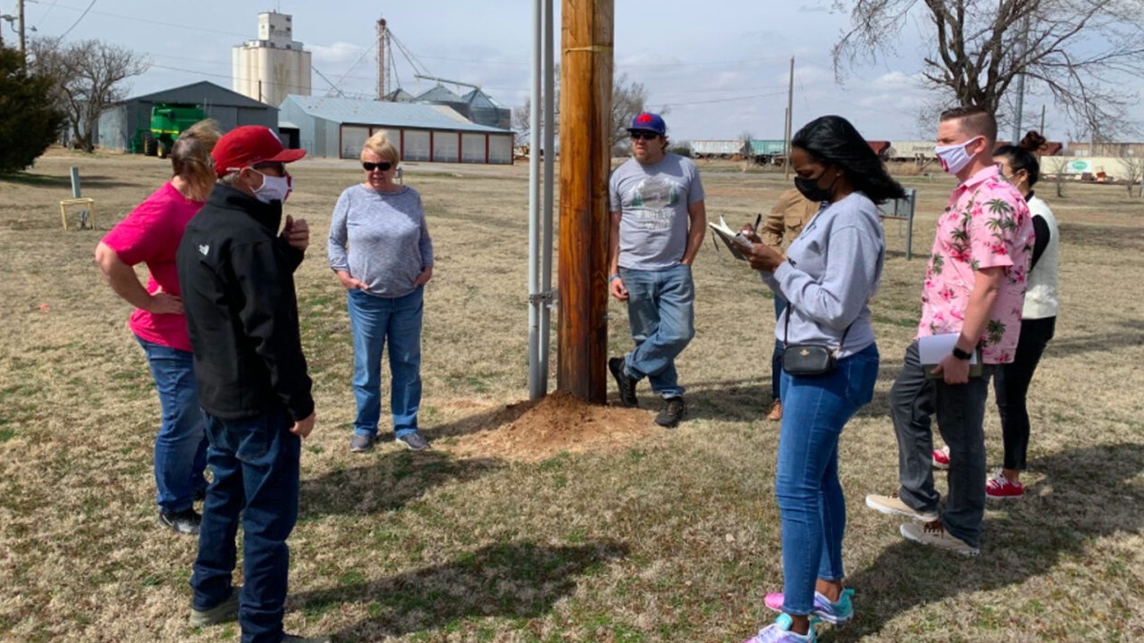 Group of people wearing masks gathered outdoors around a utility pole, taking notes and discussing the site.