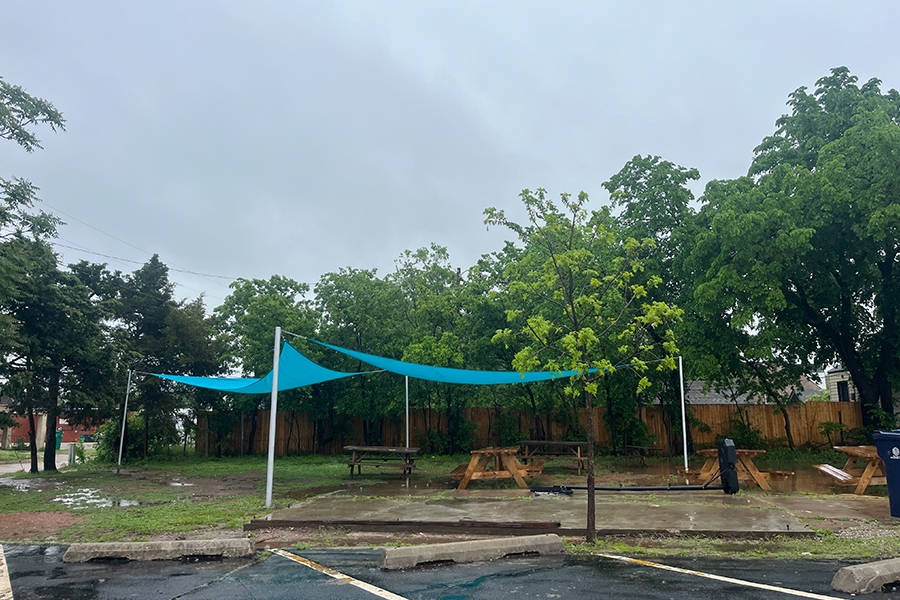 A view of the completed pavilion at Sisu Youth Services in Oklahoma City, featuring teal shade canopies stretched between metal posts over picnic tables and a concrete patio, surrounded by trees and wet pavement after rainfall.