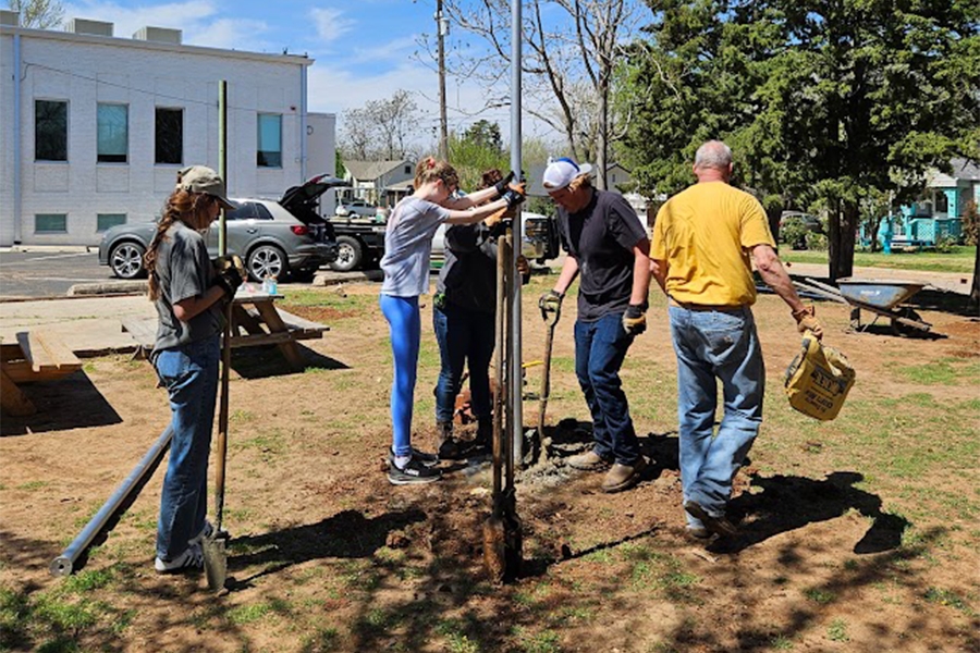 A view of the completed pavilion at Sisu Youth Services in Oklahoma City, featuring teal shade canopies stretched between metal posts over picnic tables and a concrete patio, surrounded by trees and wet pavement after rainfall.