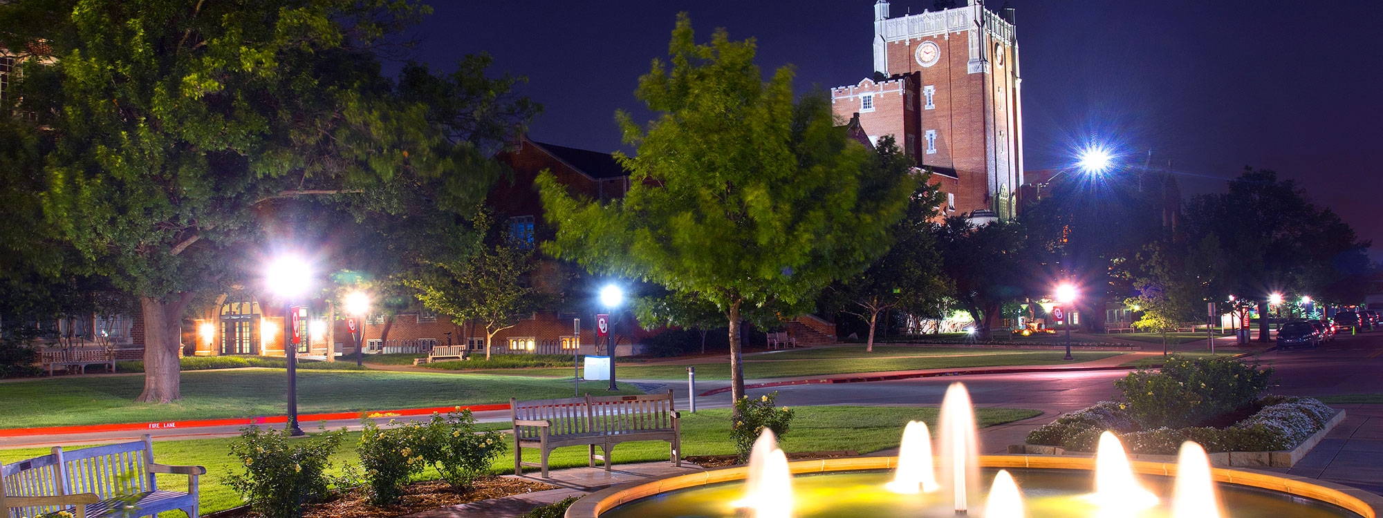 The Oklahoma Memorial Union at night, with trees, benches, and a fountain in the foreground.
