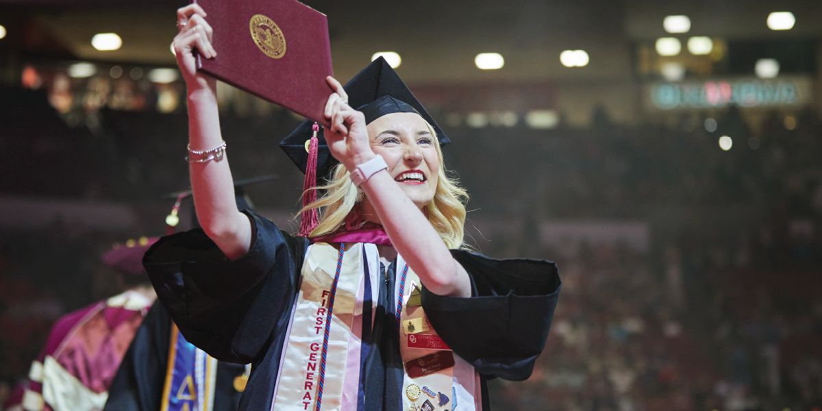 Female First-Generation graduate in cap and gown holding a diploma cover at OU's commencement ceremony. 