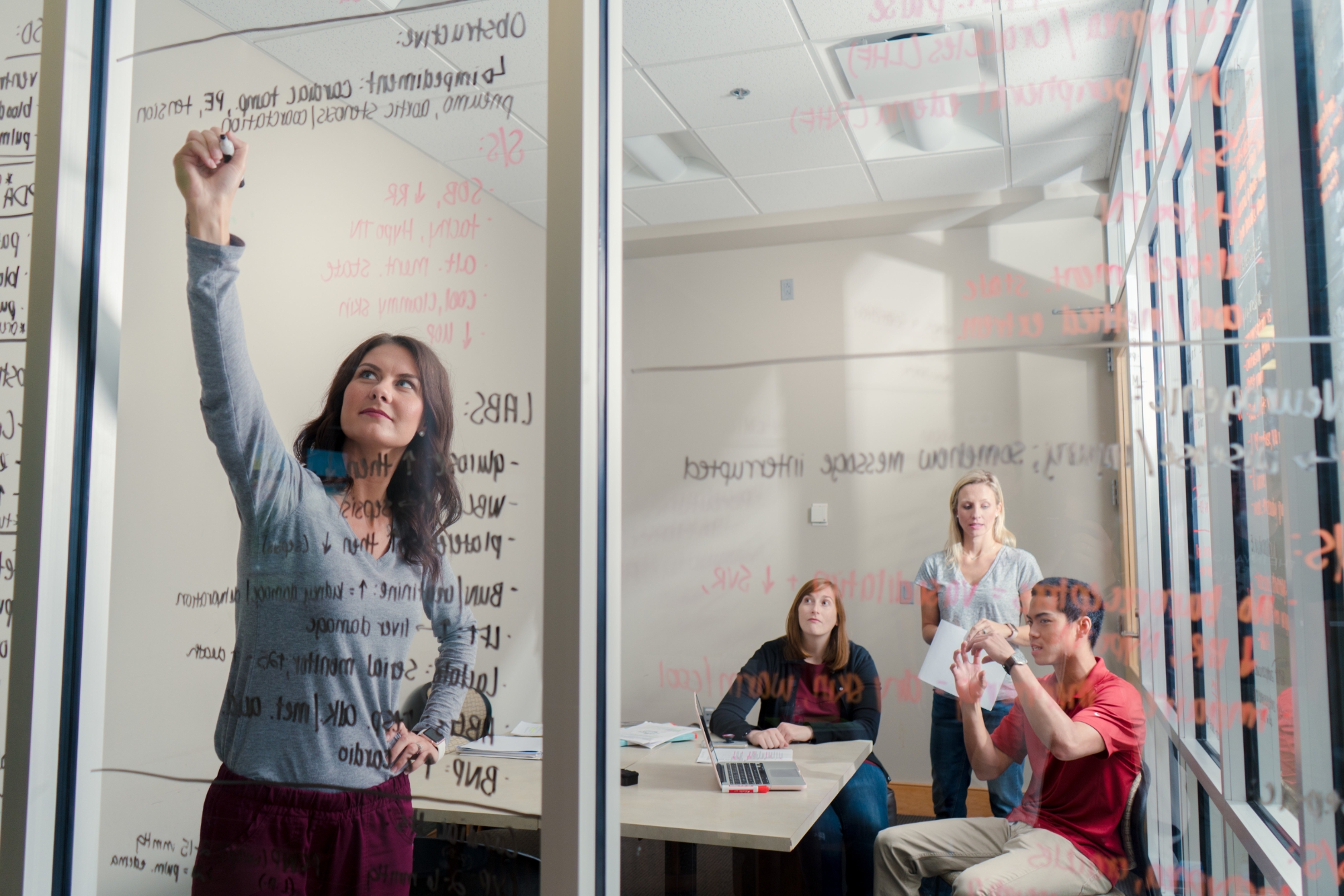 Group of students in a meeting room writing on a glass wall with expo markers
