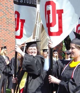 OU students in graduation regalia carrying flags with the names of different colleges .
