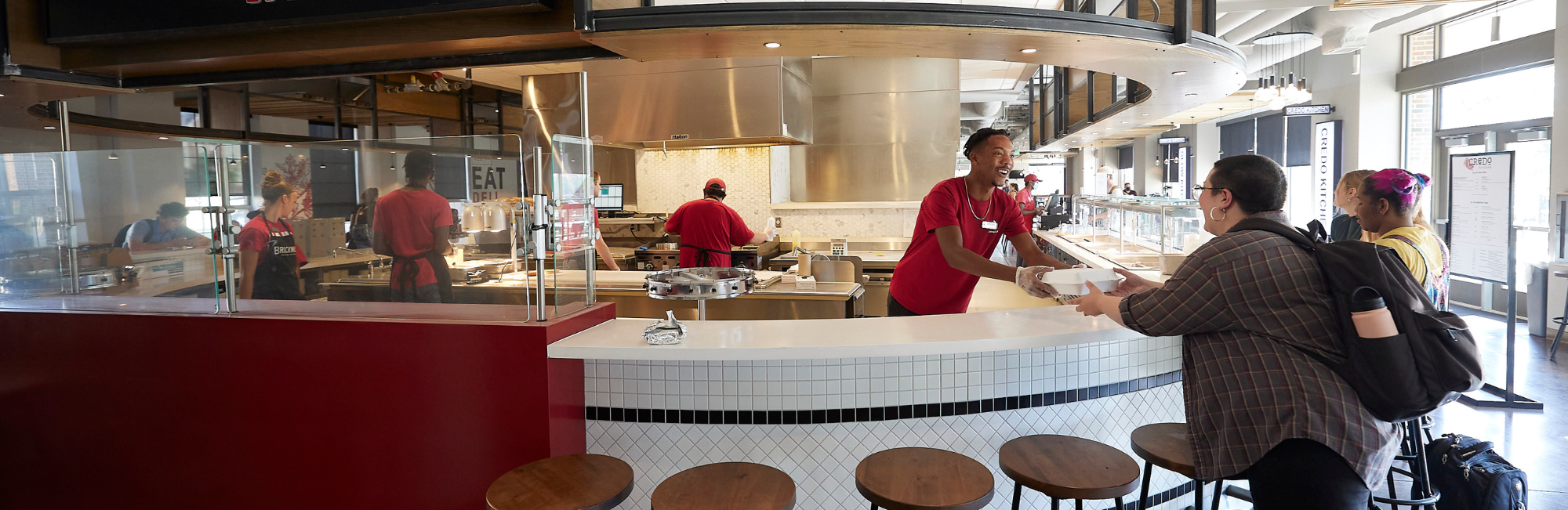 Employee in a modern dining space hands a to-go box to a customer. Other customers are sitting at a counter waiting for their order. A large menu for Credo Kitchen can be seen in the background.
