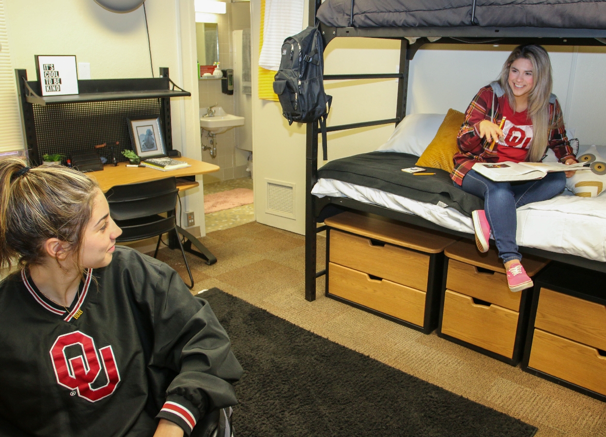 One female student sits at a desk in Couch Center and talks to her roommate who is studying on a bed.