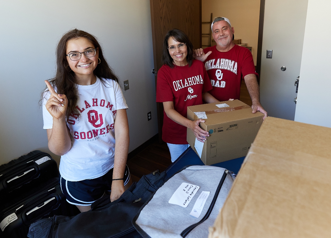 A female student stands with boxes in her Cross Village room with her two parents. T