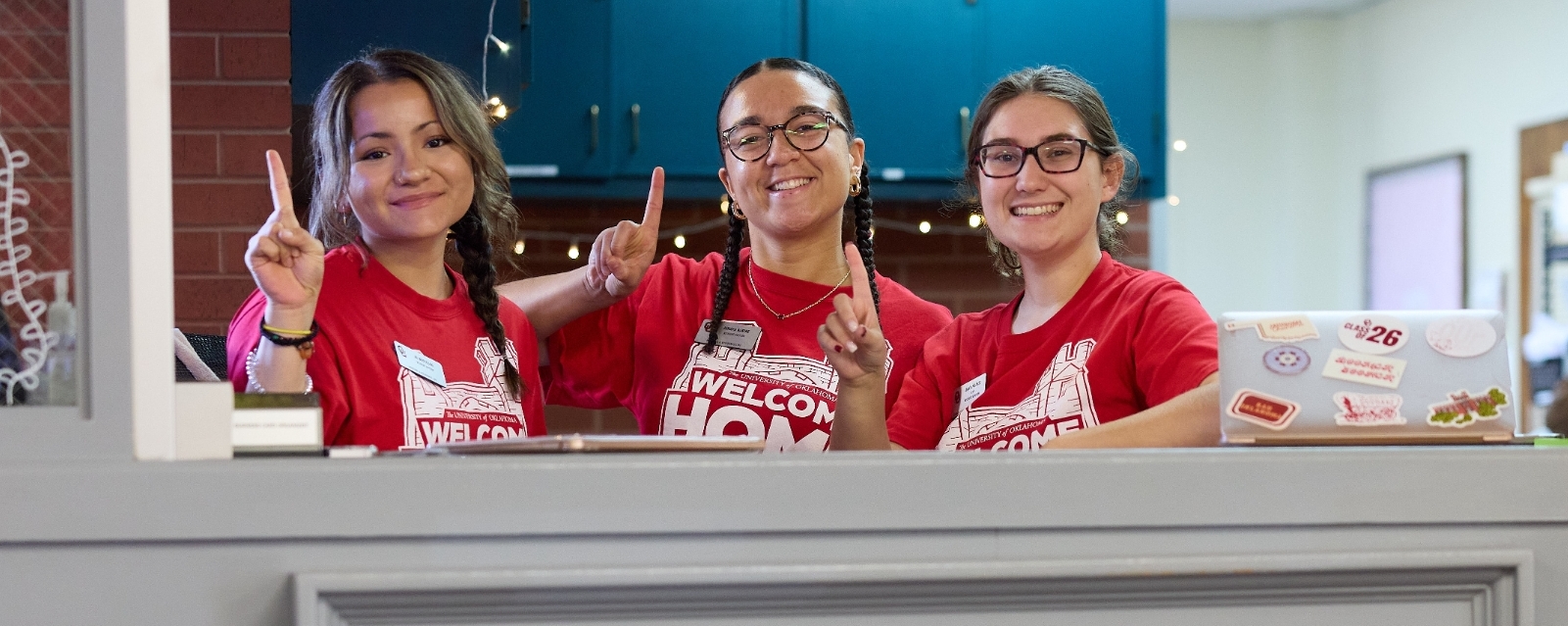 Three RAs sitting at the front desk in Walker Center while smiling at the camera. All three have red "Welcome Home" t-shirts on.
