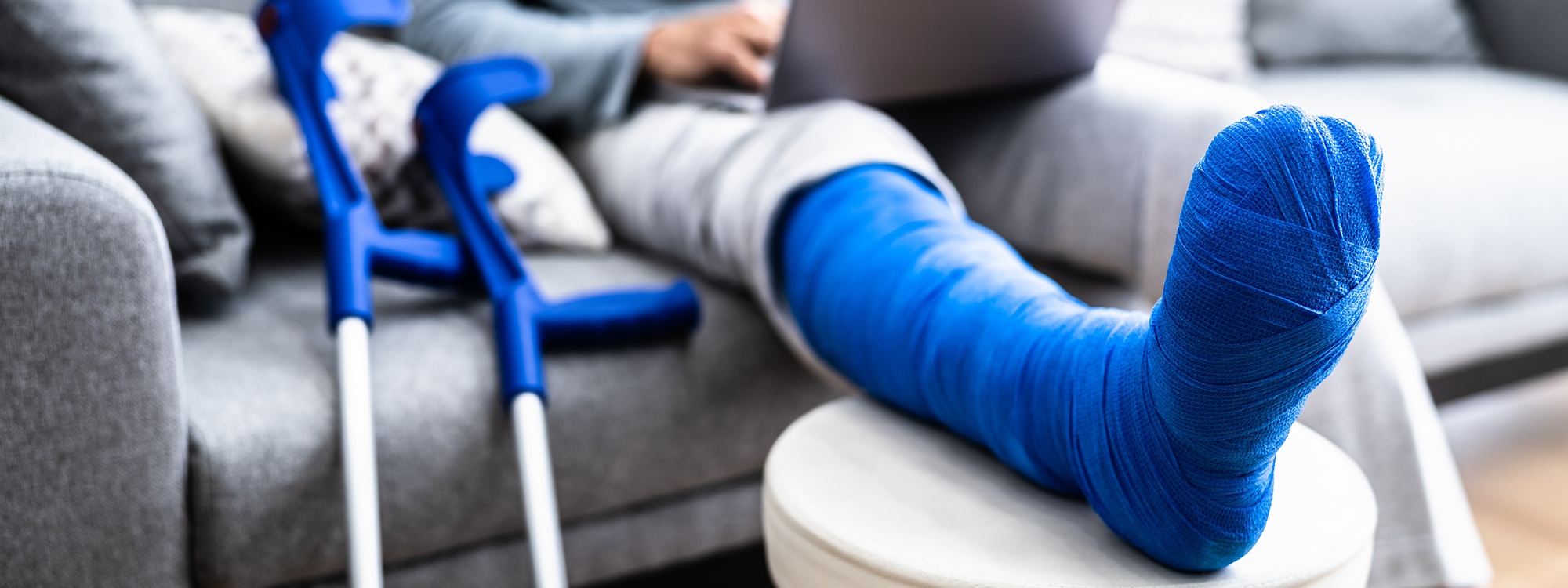 A leg in a cast resting on an ottoman while a person uses their laptop on the couch.