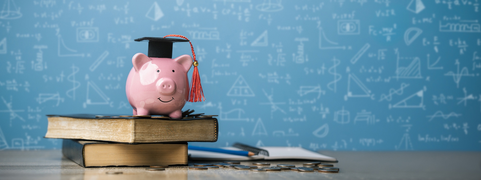Piggy bank with a graduation cap standing on top of some books and a pile of coins on a desk.