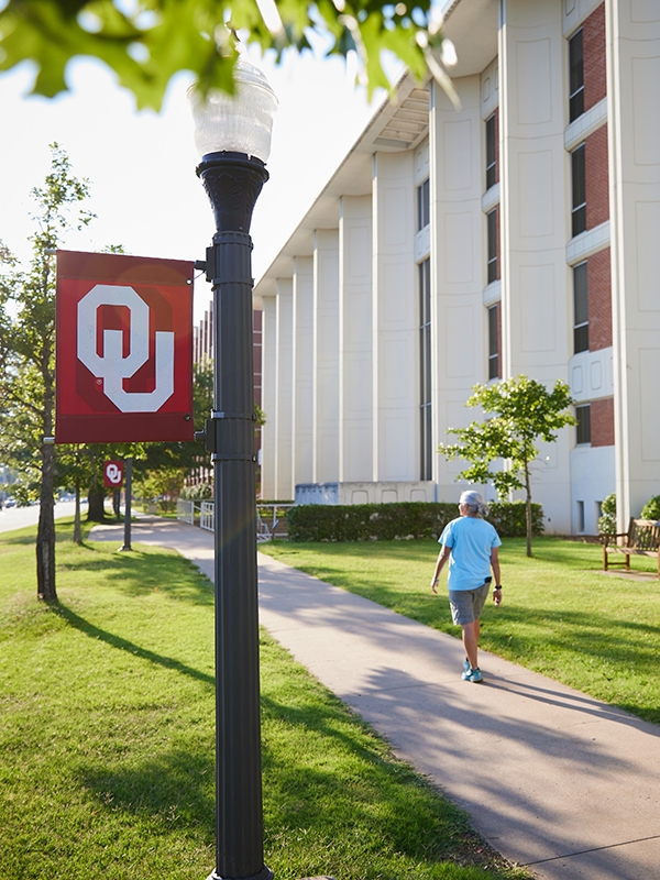 Person walking down the sidewalk next to the engineering building with a lightpost and O-U sign in the foreground.