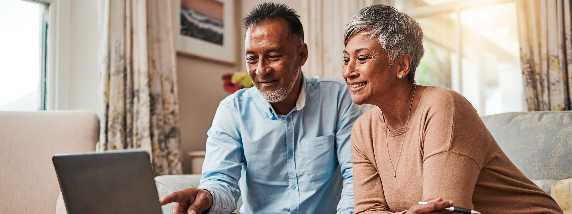An elderly couple looking and pointing at their laptop while sitting on their living room couch.