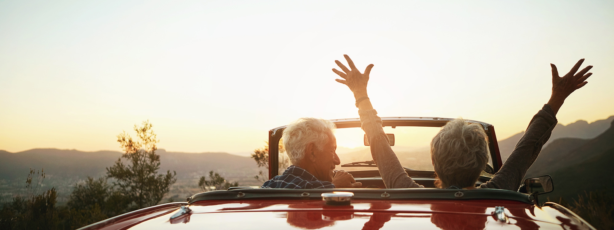 Retired couple driving into the sunset with the woman's arms in the air celebrating.