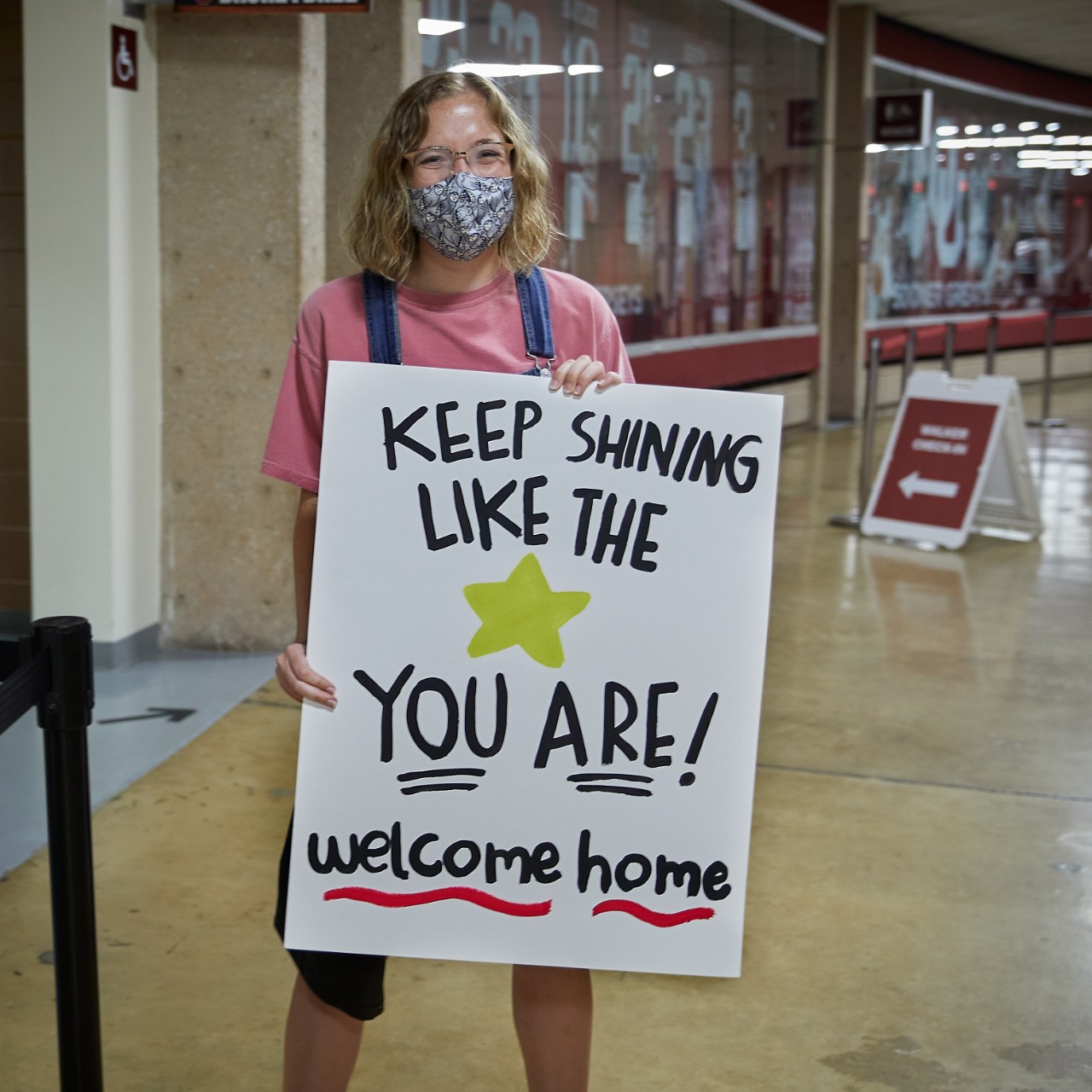 Masked student holds up a hand made sign reading "keep shining like the star you are! welcome home"