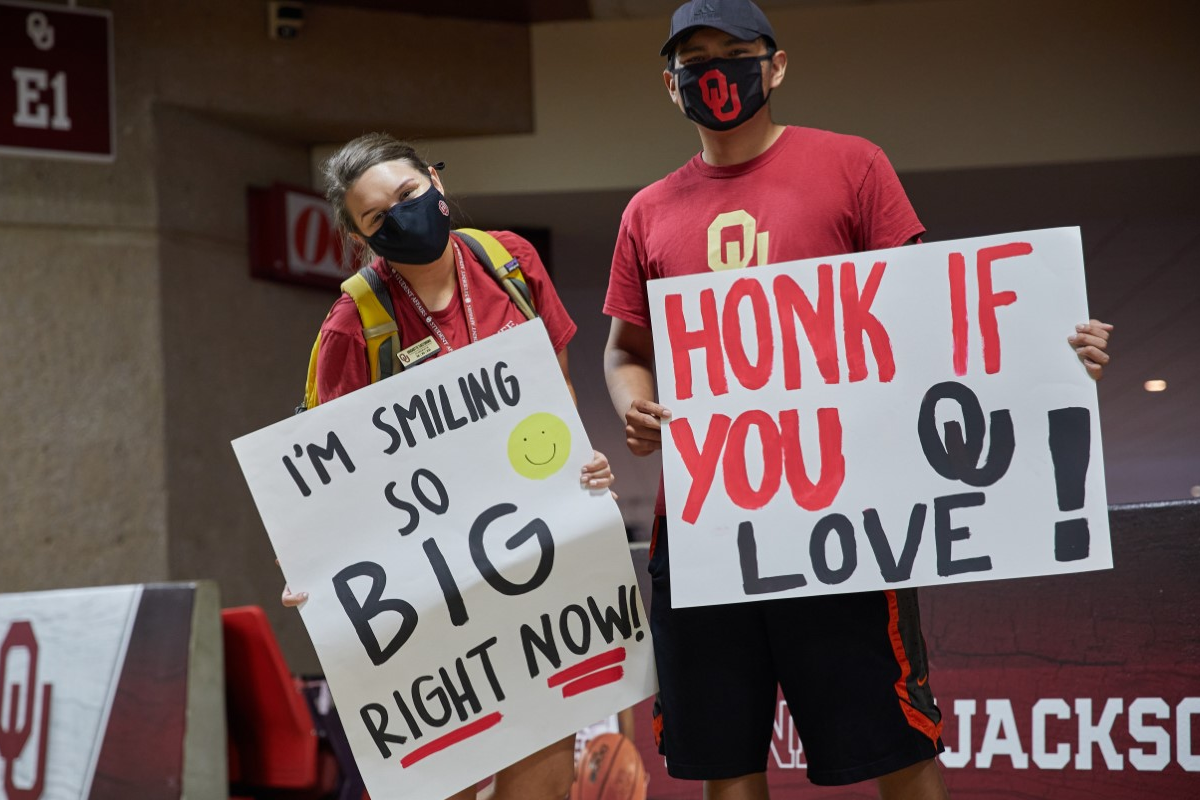 Masked students hold up hand made signs reading "Honk if you love OU!" and "I'm smiling so BIG right now!"
