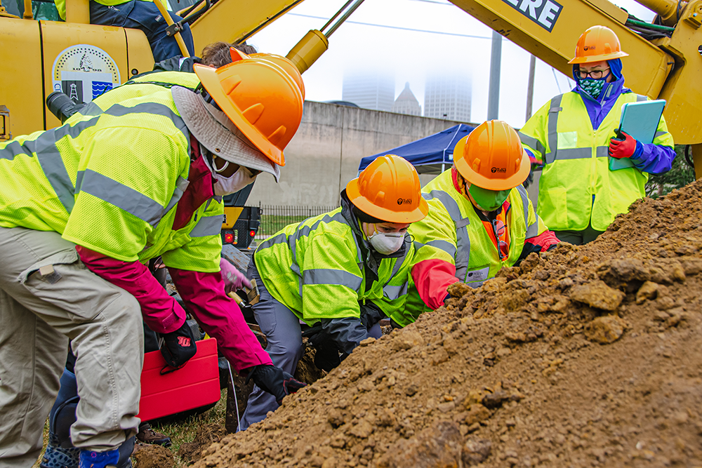 Five masked, hard-hatted workers wearing high-vis vests excavate a section of earth.
