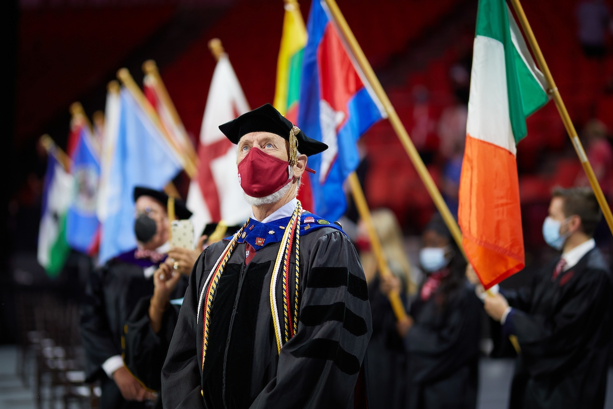 faculty member in regalia stands in front of line of international flags
