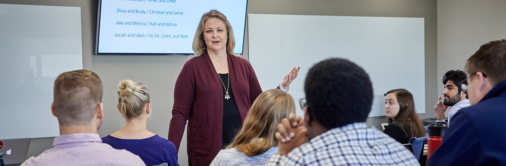 A professor speaking to a group of students, with a TV displaying group assignments.