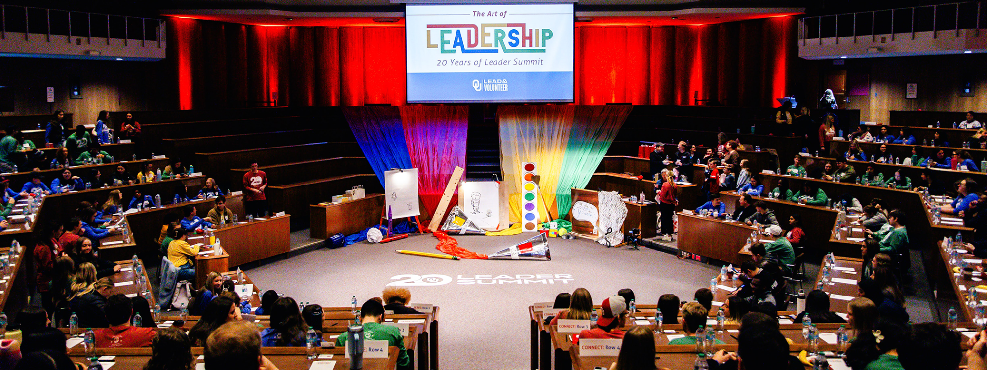 A wide shot photo at the 2025 Leader Summit. A very color filled presentation room with students in seats and a projection screen displaying "Leader Summit 2025."