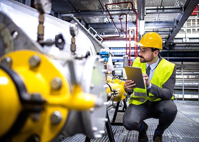 A man in a hardhat and vest, squatting and typing on a tablet.