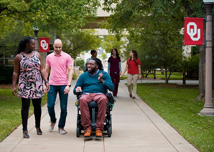 Student in a wheelchair talking with two other students on a shaded pathway on campus lined with O U flags.