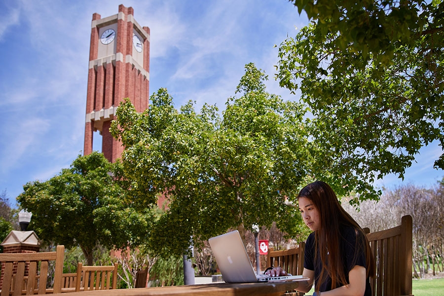 A student sitting at an outdoor table near the Bizzelle library, working on a laptop.