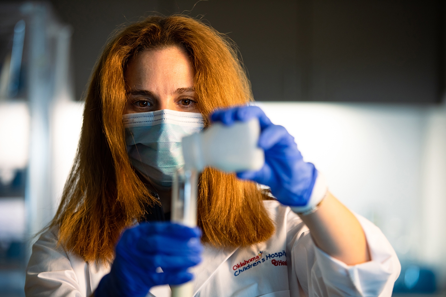 A doctor wearing a blue mask pours a chemical into a vial. 