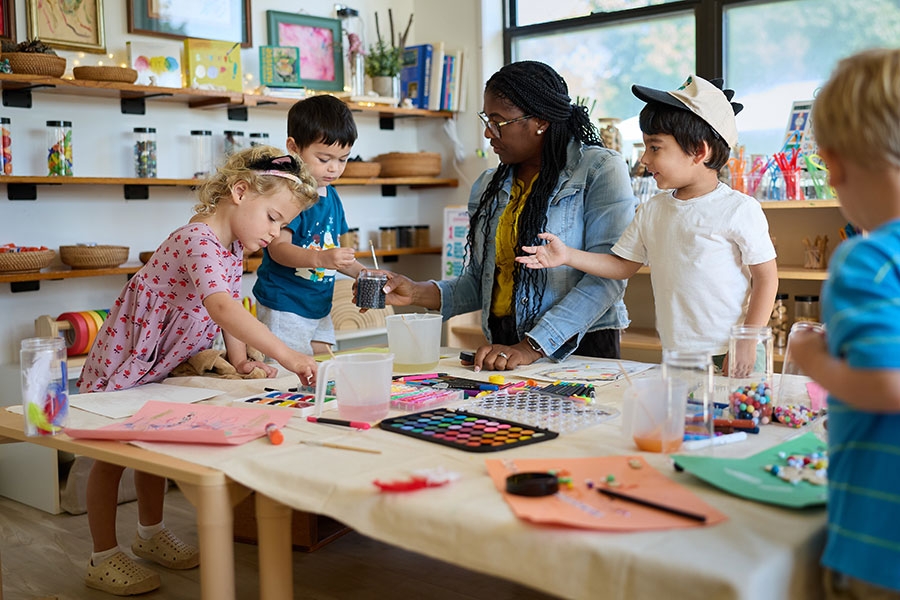 A teacher kneels at a table surrounded by children. The table is covered in arts and crafts materials.