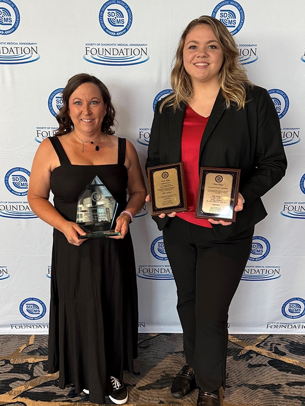 Photo of two women holding awards in front of event backdrop banner.