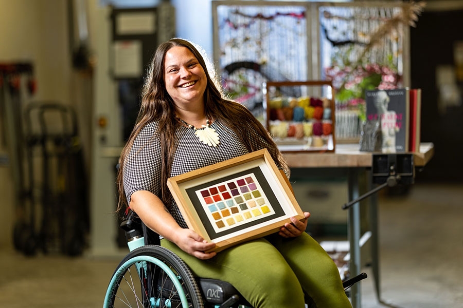A woman seated in a wheel chair holds up a piece of art in a frame.