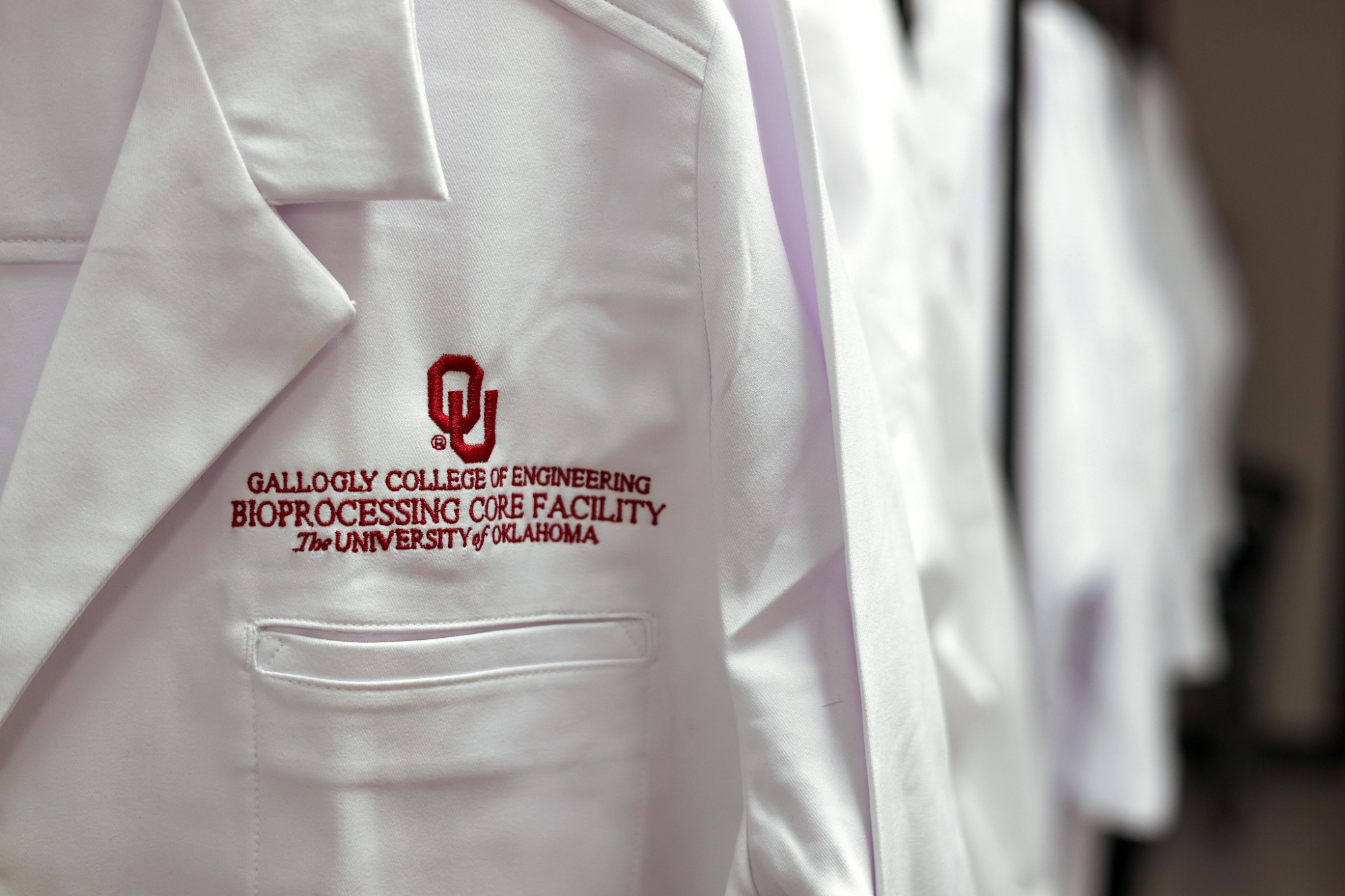 A close-up photo of a lab coat with "Gallogly College of Engineering Bioprocessing Core Facility The University of Oklahoma" embroidered on it.