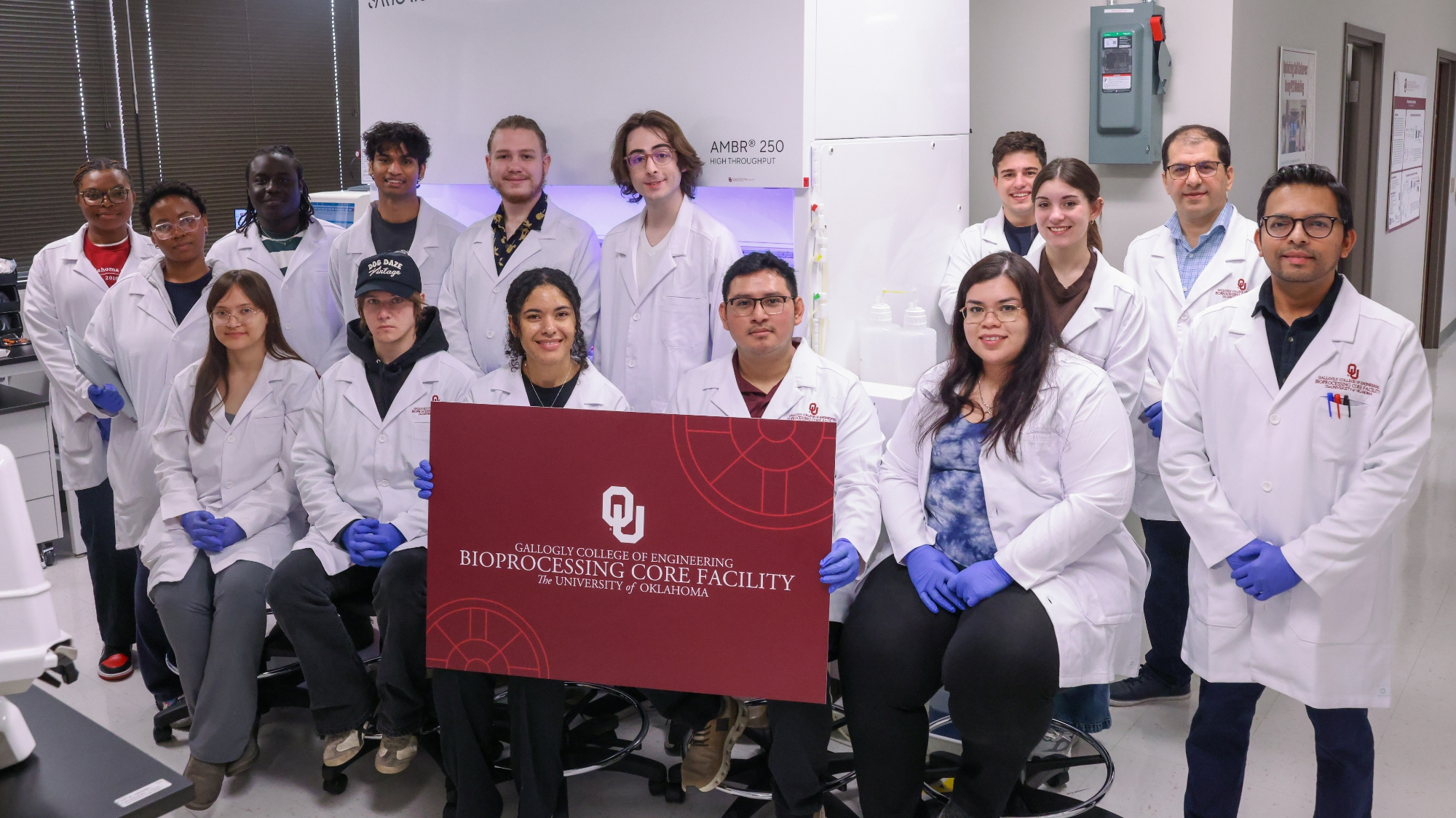 A group of people in white lab coats sitting in a lab, holding up a sign for the bioprocessing core facility.