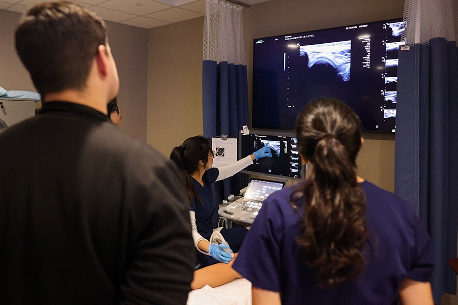 A woman in a dark blue lab coat points at an ultrasound image in front of onlookers.