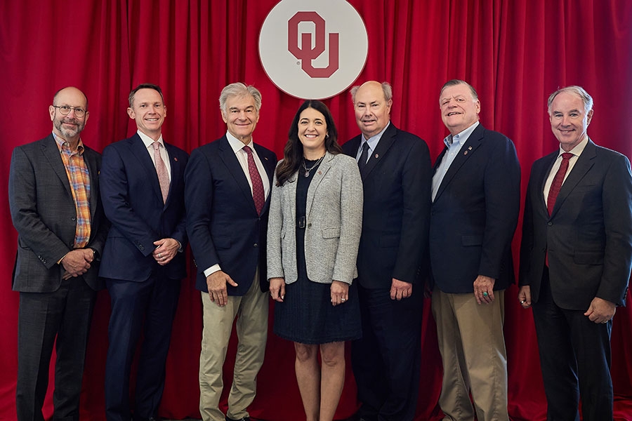 A group of six people standing in front of a red background with an OU crest on it. 