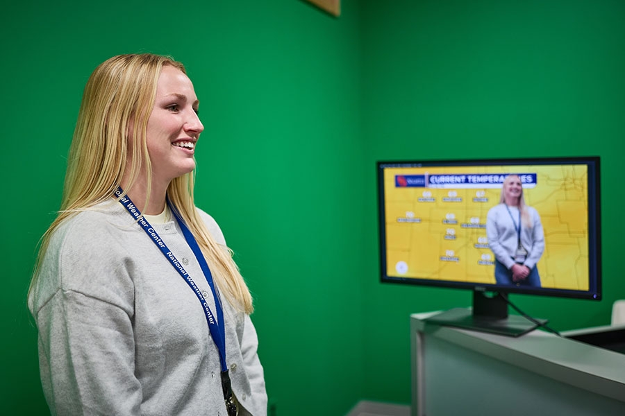 A student stands in front of a green screen. Behind her, a monitor shows her standing before a weather map of Oklahoma.