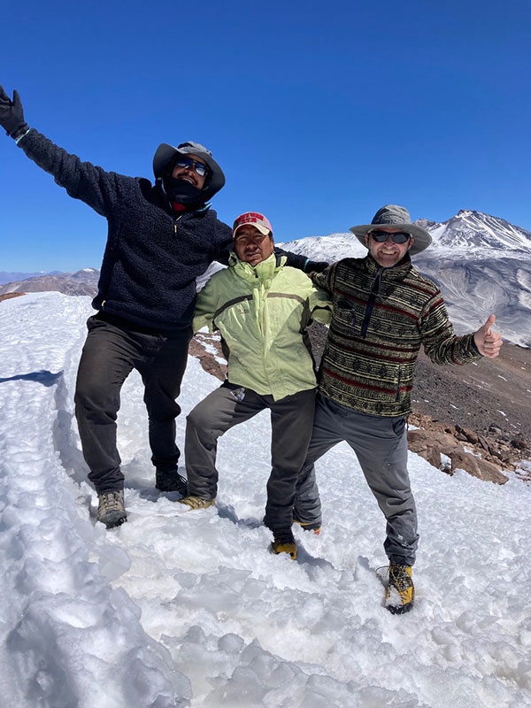 Three men in cold-weather outfits pose together on a snow-covered mountain.