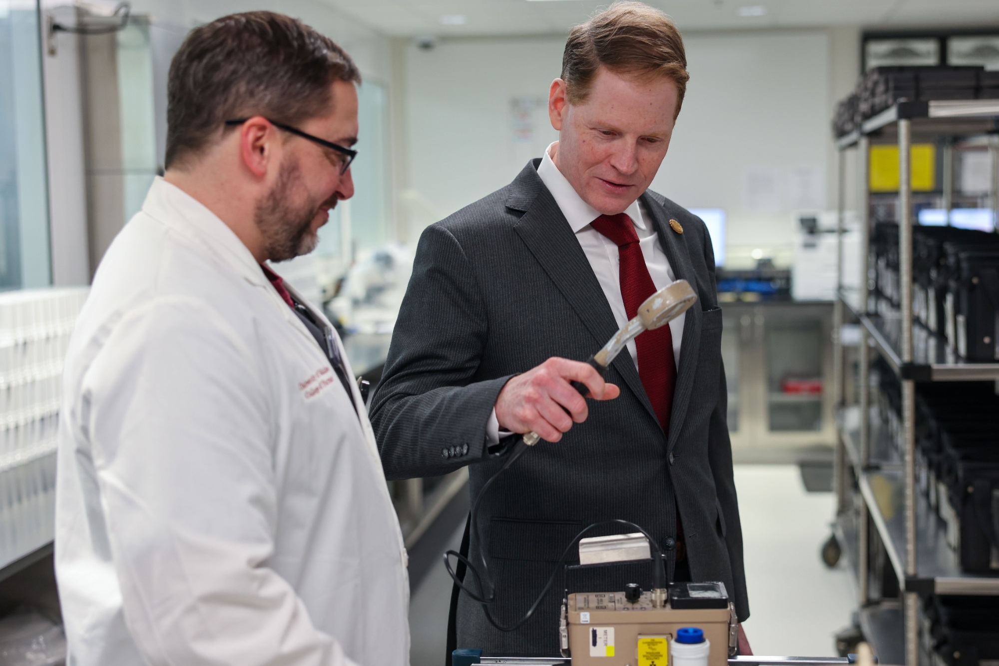 A doctor in a white coat and a man in a grey suit examine medical technology.