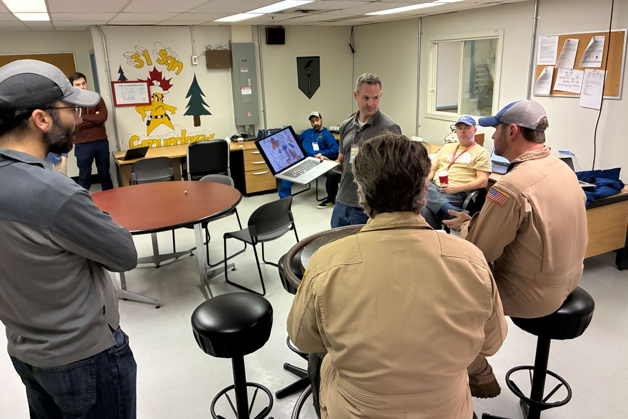 A group of people sit in a circle surrounding one individual who holds a laptop. 