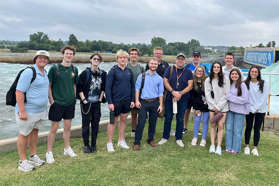 A group of mostly younger adults stands in front of a flowing river. A sign labeled Riversport Rapids hangs above part of the river.