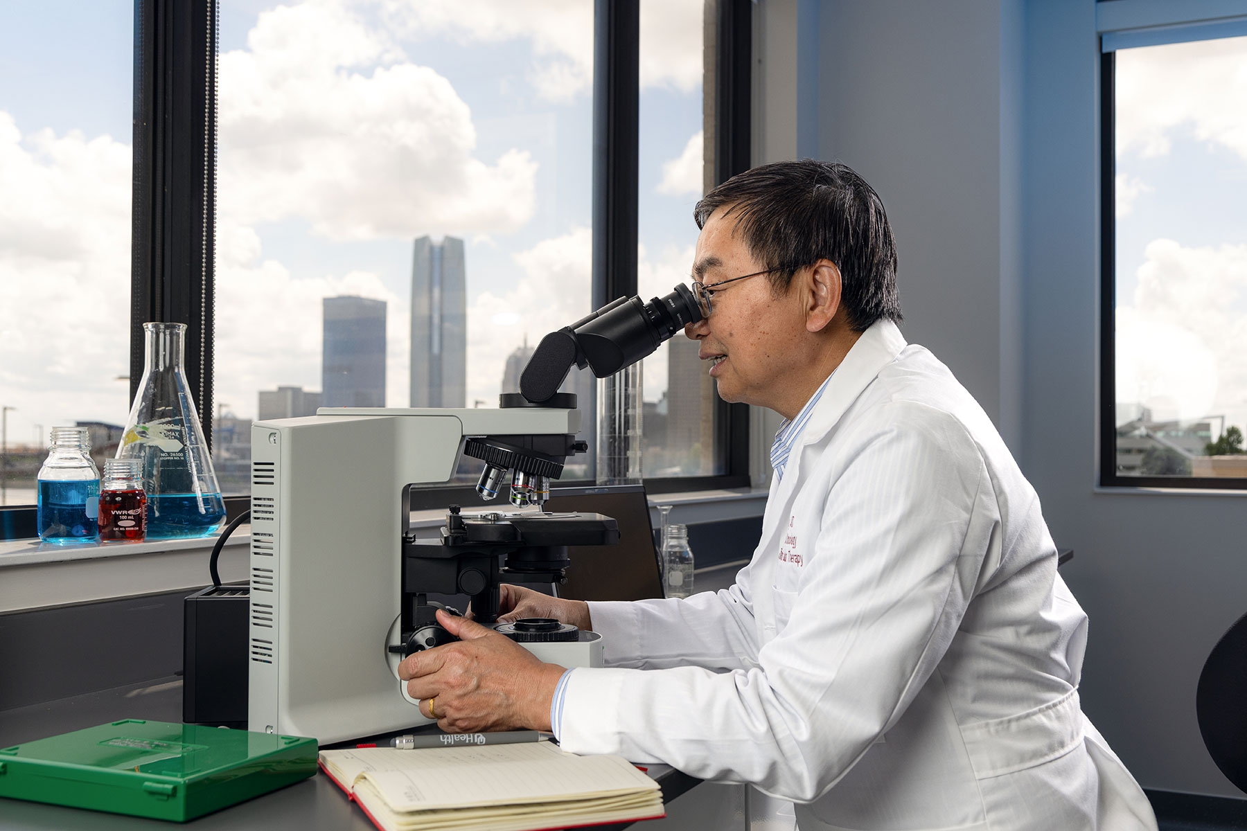 Yubin Kang looks into a microscope at a desk seated in front of a window. Outside the window is the Oklahoma City skyline.