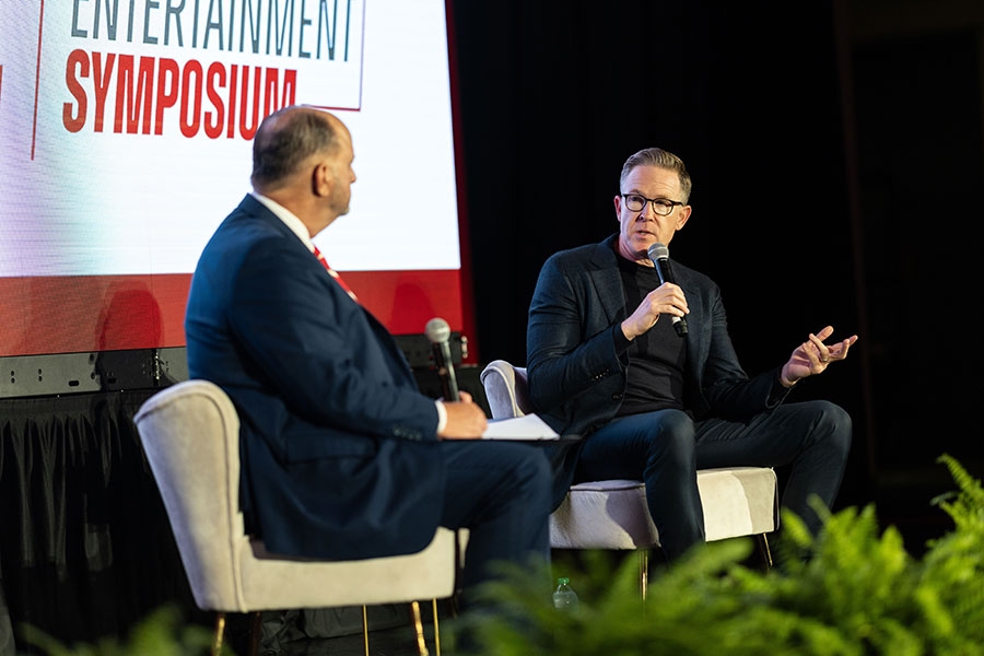 A man holding a microphone talks to another man while the two sit in chairs on a stage, during a symopsium.