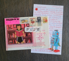 On the right of the picture is a handwritten letter in red ink, with a robot on the corner of the paper. On top of the letter is a colorful envelope with a cartoon woman reading a book and a "Via Air Mail Par Avion" sticker and a four stamps.