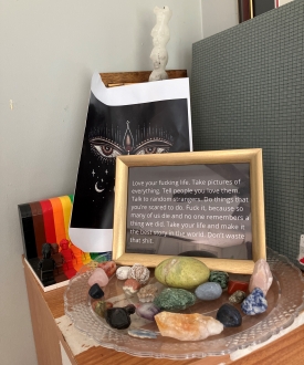 A framed photo of text propped up on a clear glass plate and surrounded by colorful rocks. The framed image reads, "Love your fucking life. Take pictures of everything. Tell people you love them. Talk to random strangers. Do things that you're scared to do. Fuck it, because so many of us die and no one remembers a thing we did. Take your life and make it the best story in the world. Dont' waste that shit."