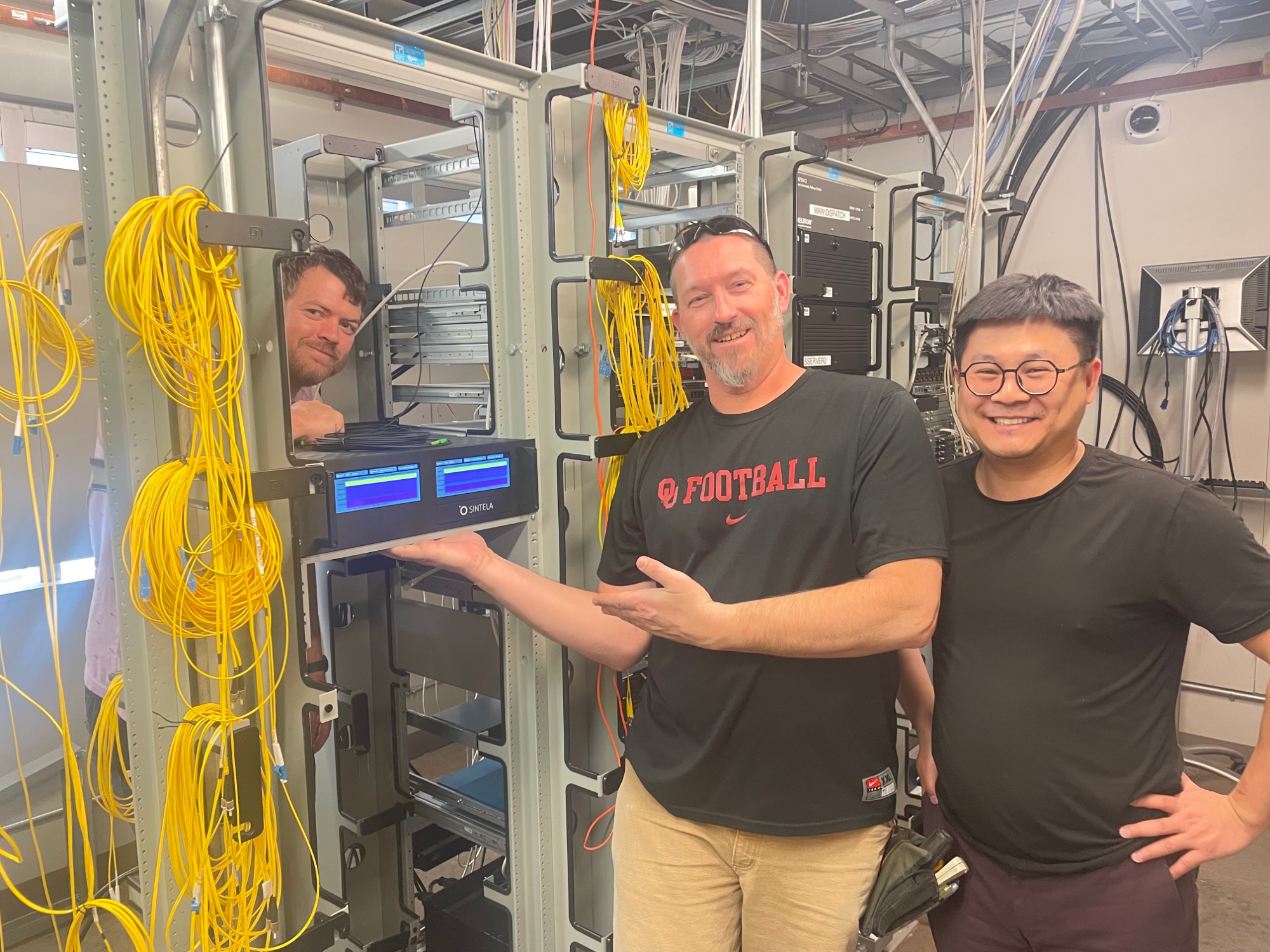Three men smile while standing next to a tower of technology equipment.