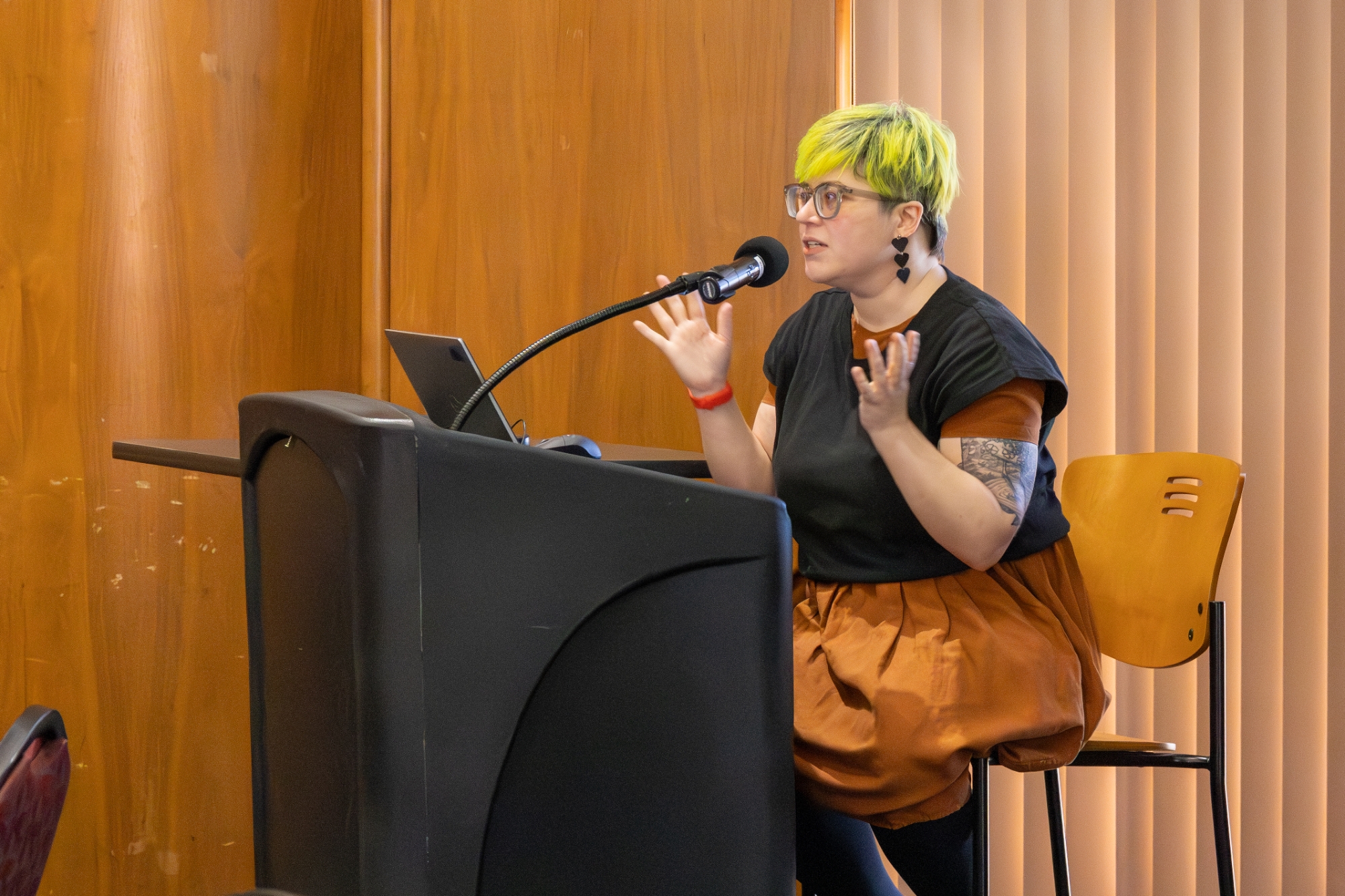 Person sitting on a tall chair, talking at a podium.