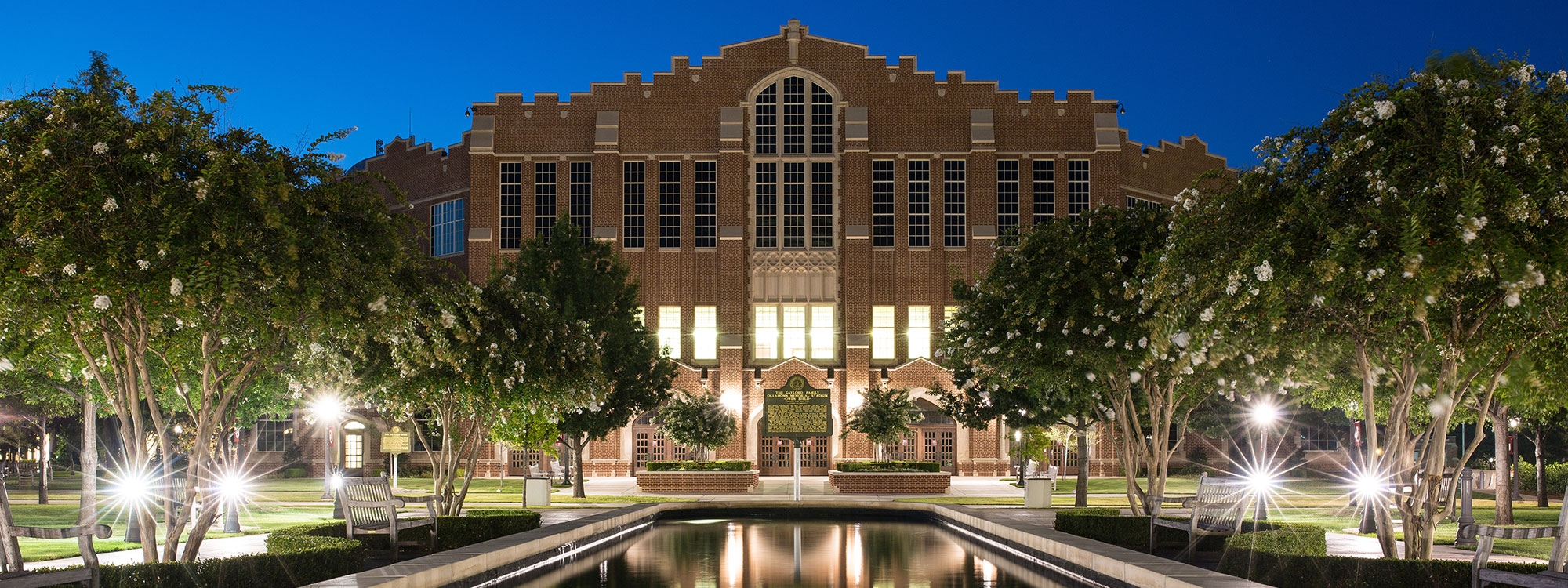The McCasland Field House in the evening, with a water feature and trees in the foreground.