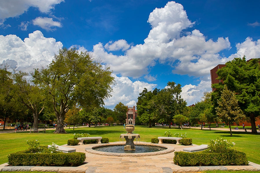 A water fountain in the foreground, with greenery and blue sky with white clouds in the background.