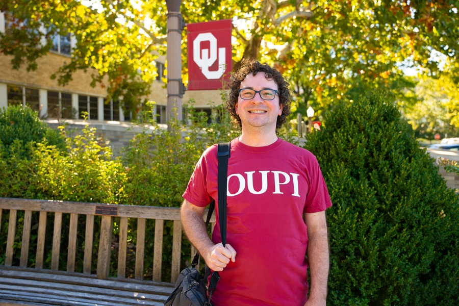 Student wearing OUPI shirt standing on campus in front of a bench and an OU flag.