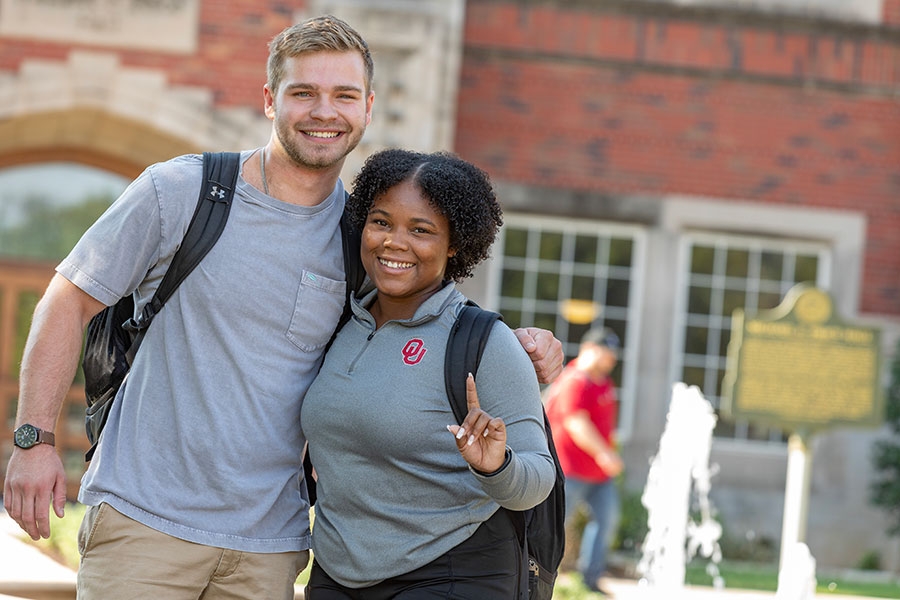 Two smiling students with backpacks standing in from of  the Fountain outside of Price Hall. 
